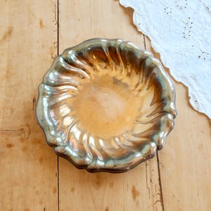 May include: A round, shallow bowl with a metallic, iridescent finish that shifts between gold, bronze, and dark blue tones. The bowl features a distinct, swirling, fluted pattern along its rim, creating a textured, wavy edge that catches light.