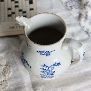 Puede incluir: Una jarra de cer&aacute;mica blanca con dise&ntilde;os florales y de p&aacute;jaros azules. La jarra tiene un pico y un asa. Un libro de crucigramas est&aacute; en el fondo. La jarra est&aacute; sobre un pa&ntilde;o blanco.