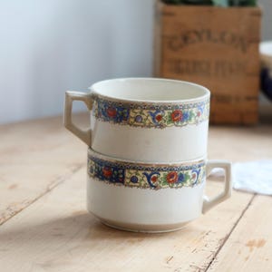 May include: Two stacked cream-coloured ceramic cups featuring a decorative band around the upper rim. The pattern consists of blue, red, and yellow floral motifs with intricate gold-toned detailing. Each cup has a geometric, angular handle.