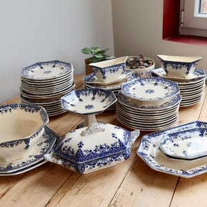 Peut inclure: Une grande collection de vaisselle en céramique couleur crème ornée d'un motif floral bleu complexe. L'ensemble comprend des assiettes octogonales empilées, une soupière avec couvercle, un bol sur pied, une saucière et des plats de service.