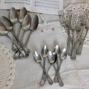 Pode incluir: Um conjunto de colheres e garfos de prata com alças ornamentadas. As colheres e os garfos estão dispostos sobre uma toalha de mesa de linho branco com dois guardanapos de crochê.