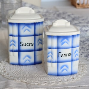 Puede incluir: Dos botes de cerámica blancos con patrones de cuadros azules y tapas blancas. Uno dice "Sucre" y el otro "Farine" en texto negro. Los botes están sobre un mantel individual de encaje blanco.