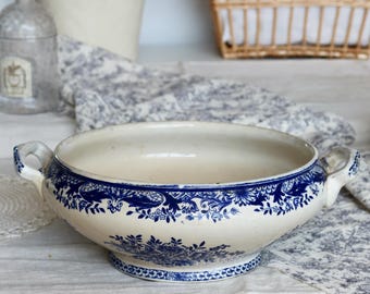 Op de afbeelding: Een witte keramische serveerschaal met een blauw bloemmotief rond de rand en de basis. De schaal heeft twee handvatten en staat op een kleine voet. Een decoratief stuk voor het serveren van voedsel.