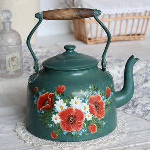 Puede incluir: Una tetera vintage de color verde azulado con un asa de madera y un diseño floral con amapolas rojas, margaritas blancas y nomeolvides azules. La tetera está sobre un tapete de encaje blanco, evocando una estética rústica.
