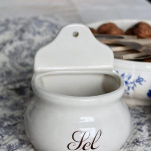 Può includere: Una saliera in ceramica bianca con corpo curvo e parte superiore rettangolare con foro per appenderla. La parola "Sel" è scritta in elegante corsivo sul davanti. Lo sfondo comprende una tovaglia fantasia e altri utensili da cucina.