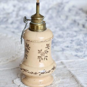 May include: Cream-colored antique oil lamp with a brass top and a wick. The lamp features floral designs in brown ink. The lamp is sitting on a white lace doily. A chain is attached to the brass top.