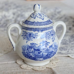 May include: A white and blue ceramic sugar bowl with a lid. The bowl features a blue pastoral scene and floral patterns. It has two handles and a small knob on the lid. The bowl is sitting on a white lace doily.