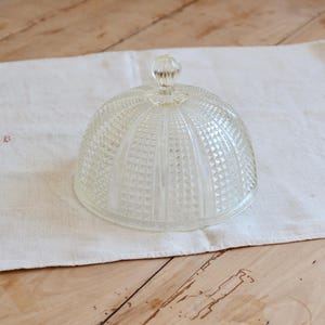 Peut inclure: Une cloche en verre transparent avec un motif géométrique texturé. Le couvercle en forme de dôme a un petit bouton rond sur le dessus. La cloche repose sur un tissu blanc, avec une surface en bois en arrière-plan. Cet article est adapté pour exposer des pâtisseries.