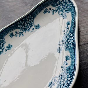 May include: White ceramic serving dish with a blue floral pattern around the rim. The dish has a unique, curved shape and is set against a dark wooden surface. The blue design includes flowers and geometric shapes.