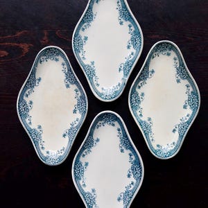 May include: Four white, diamond-shaped serving dishes with a blue floral border. The dishes are arranged on a dark wooden surface, showcasing their vintage design. Each dish has a slightly off-white center.