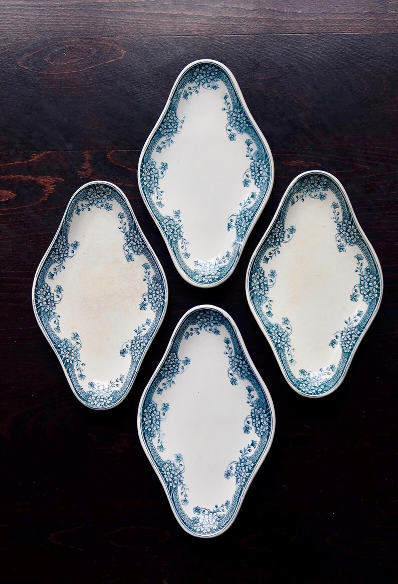 May include: Four white, diamond-shaped serving dishes with a blue floral border. The dishes are arranged on a dark wooden surface, showcasing their vintage design. Each dish has a slightly off-white center.