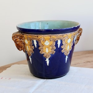 Peut inclure: Un pot de fleurs en céramique bleu avec des détails dorés et blancs ornés. Le pot présente des poignées en tête de lion et un intérieur bleu clair. Cette pièce décorative convient à l'intérieur ou à l'extérieur.