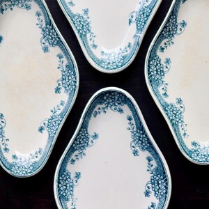 May include: Four white, oval-shaped plates with a teal floral design around the edges. The plates are arranged on a dark wooden surface, showcasing a vintage aesthetic. The floral pattern adds a touch of elegance.