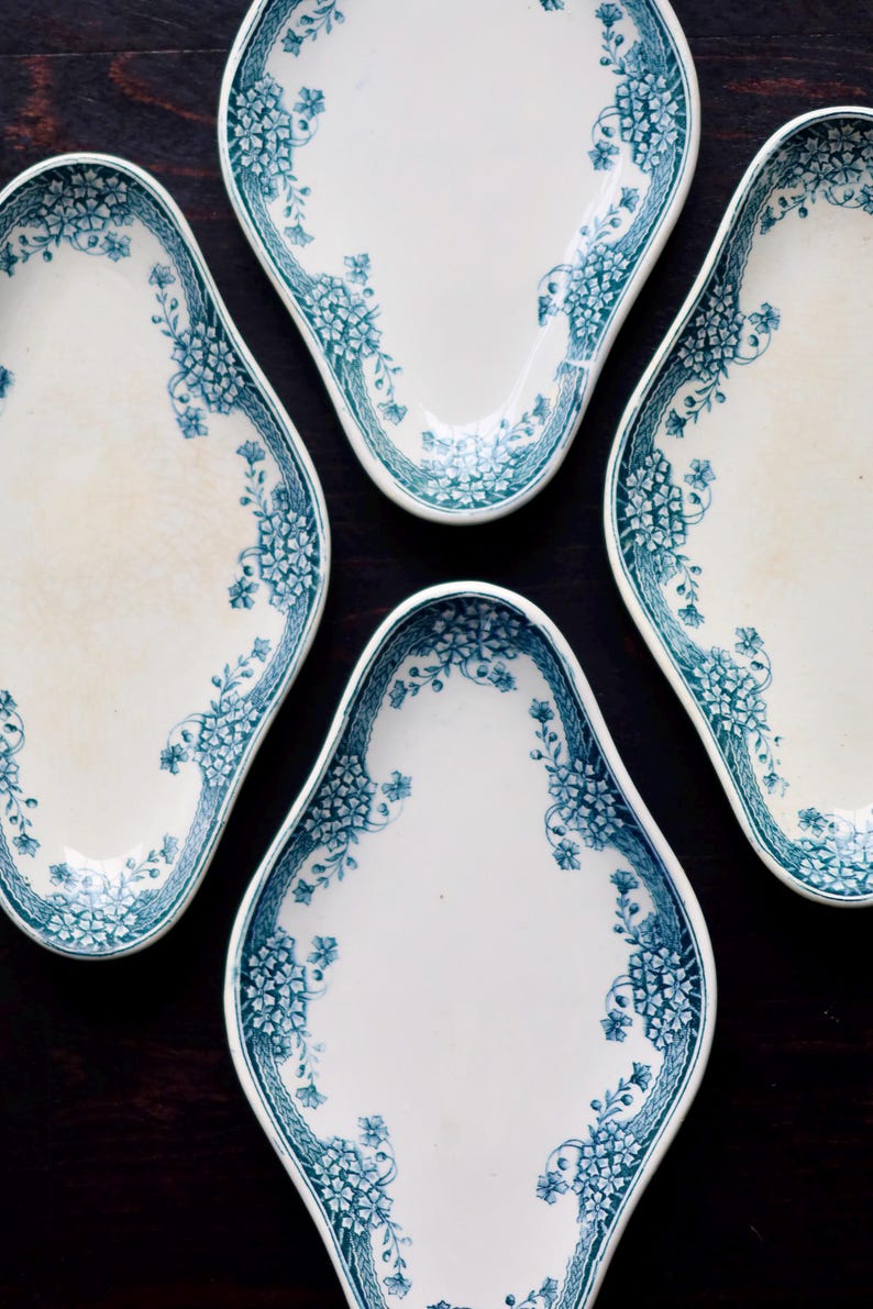 May include: Four white, oval-shaped plates with a teal floral design around the edges. The plates are arranged on a dark wooden surface, showcasing a vintage aesthetic. The floral pattern adds a touch of elegance.