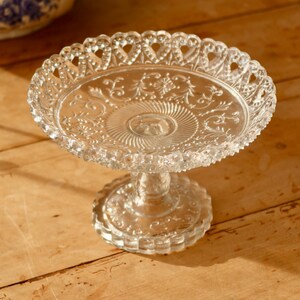 May include: A clear glass cake stand with a decorative, scalloped edge and intricate floral pattern. The stand has a pedestal base.