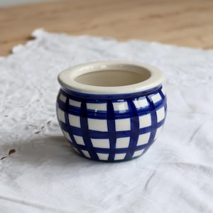 Może przedstawiać: Mała, okrągła ceramiczna miseczka z błyszczącym wykończeniem w ciemnoniebiesko-białą kratkę. Misa posiada szeroki, gładki, kremowy brzeg oraz głębokie, otwarte wnętrze, charakteryzujące się klasycznym, zaokrąglonym kształtem i płaską podstawą.