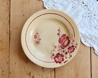 May include: A round, cream-colored ceramic plate featuring a thin, dark red circular border line and a decorative floral pattern in shades of deep red. The floral design is positioned on the right side of the plate, showcasing stylized blossoms.