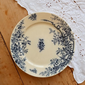 Può includere: Piatto in ceramica color crema con un motivo floreale blu. Il piatto presenta un motivo floreale centrale e un bordo con fiori stilizzati e forme geometriche. Appoggiato su una superficie di legno con un panno di pizzo bianco.