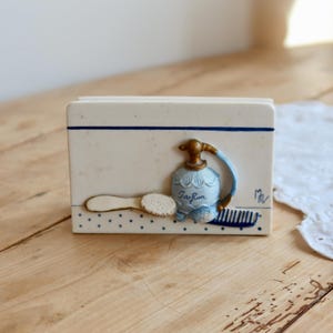 Peut inclure: Un porte-serviettes rectangulaire en céramique blanche avec une bande horizontale bleue et un motif à pois bleus à la base. L'avant présente un relief d'un flacon de parfum bleu clair étiqueté Parfum, une brosse blanche et un peigne bleu.
