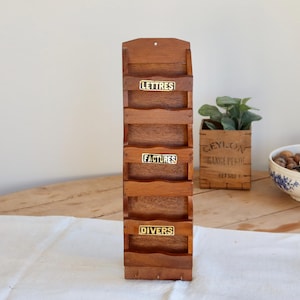 Peut inclure: Un organiseur de courrier en bois avec plusieurs compartiments étiquetés "Lettres", "Factures" et "Divers". L'organisateur est en bois aux tons chauds et a une esthétique vintage. Un petit coffret en bois avec de la verdure et un bol bleu et blanc sont également visibles.