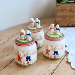 Può includere: Tre barattoli in ceramica bianca con coperchio, ciascuno sormontato da due piccole statuette di anatre con fiocchi rossi e blu. I barattoli presentano un rilievo di anatre vicino a una staccionata in legno, dipinti con tocchi di verde, rosso e giallo.
