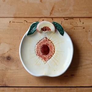 May include: A white ceramic dish shaped like a peach with green leaves. The dish has a brown center and a raised rim.