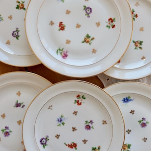 Puede incluir: Un conjunto de platos de porcelana blanca con bordes dorados. Cada plato está decorado con pequeños arreglos florales coloridos y detalles dorados. Los platos están dispuestos sobre una superficie de madera, mostrando una estética vintage.