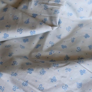 Puede incluir: Tela blanca con un patrón repetitivo de lazos azul claro, diseños florales y pequeños puntos. La tela está doblada, mostrando los detalles y la textura del patrón. El tema general sugiere ropa de cama o textil.