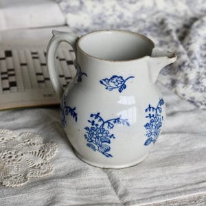 Puede incluir: Jarra de cer&aacute;mica blanca con dise&ntilde;os florales azules. La jarra tiene un asa y un pico. Un libro de crucigramas y un tapete de encaje est&aacute;n en el fondo.