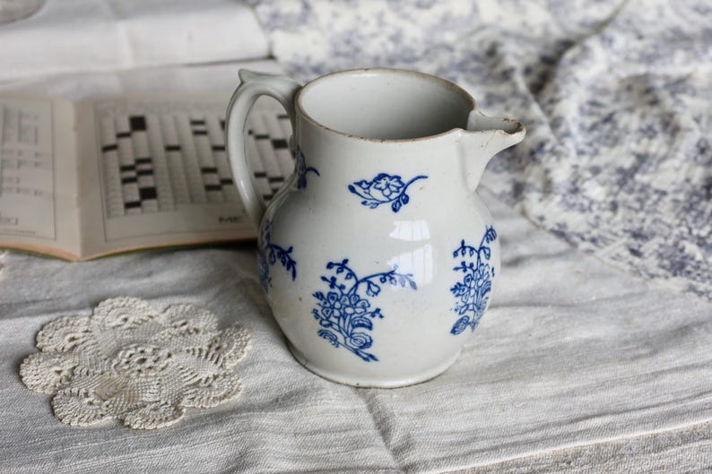 Puede incluir: Jarra de cer&aacute;mica blanca con dise&ntilde;os florales azules. La jarra tiene un asa y un pico. Un libro de crucigramas y un tapete de encaje est&aacute;n en el fondo.