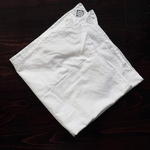 May include: A folded white linen napkin with embroidered floral and geometric details at the corner. The napkin is set against a dark brown wooden surface, creating a contrast that highlights the crisp white fabric and delicate embroidery.