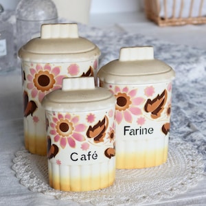 Puede incluir: Juego de tres botes de cocina de cerámica con tapas. Los botes están decorados con motivos florales rosas y marrones. El bote más pequeño tiene la palabra "Café" impresa, y el más grande "Farine". Los botes tienen una base y tapas de color crema.