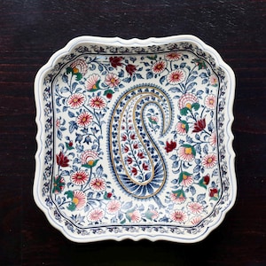 May include: A square, white ceramic serving dish with a scalloped edge. The dish is decorated with a floral pattern in red, blue, green, and yellow, and features a central paisley design.
