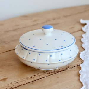 May include: A round, cream-coloured ceramic lidded dish featuring a pattern of small blue polka dots. The lid has a solid blue knob handle, and the base of the dish is designed with a pleated, ruffled edge detail around the circumference.