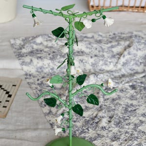 May include: A green metal jewelry display stand shaped like a flowering vine. The stand features white flower and green leaf accents. The base is round and the stand has curved arms for hanging jewelry. The stand is on a patterned surface.