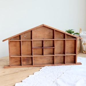 May include: A brown wooden display shelf shaped like a house with a gabled roof. The structure features multiple rectangular compartments of varying sizes arranged in rows, providing open storage space for small items within the wooden frame.