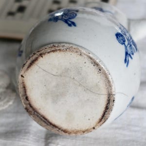 Puede incluir: Jarra de cer&aacute;mica blanca con dise&ntilde;os florales azules. La base de la jarra muestra signos de antig&uuml;edad con un borde marr&oacute;n oscuro y una grieta. La jarra est&aacute; sobre una superficie blanca.