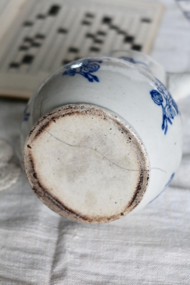 Puede incluir: Jarra de cer&aacute;mica blanca con dise&ntilde;os florales azules. La base de la jarra muestra signos de antig&uuml;edad con un borde marr&oacute;n oscuro y una grieta. La jarra est&aacute; sobre una superficie blanca.