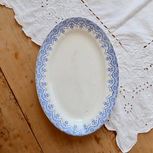 May include: An oval, white ceramic serving platter with a blue floral border. The platter is set on a wooden surface next to a white, embroidered cloth. The blue border features a repeating pattern of stylized flowers and geometric shapes.