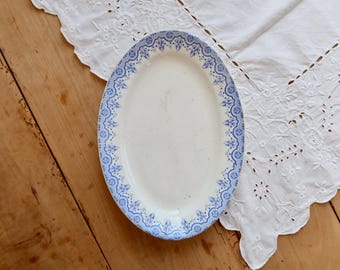 May include: An oval, white ceramic serving platter with a blue floral border. The platter is set on a wooden surface next to a white, embroidered cloth. The blue border features a repeating pattern of stylized flowers and geometric shapes.