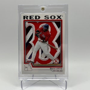 May include: A baseball card featuring David Ortiz, a Red Sox player, wearing a red and white uniform. The card has a red, white, and blue background with a large number 34 on it. The card is in a plastic protective case.