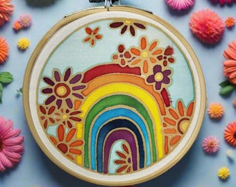 Puede incluir: Un bastidor de bordado colorido con un diseño de arcoíris y flores. El arcoíris está compuesto por arcos rojos, naranjas, amarillos, verdes, azules y morados. Las flores son en tonos naranja, morado y amarillo. El bastidor está rodeado de flores coloridas.