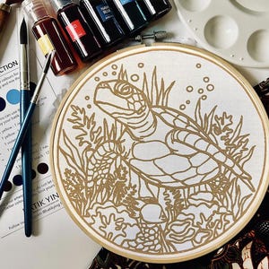 May include: A white embroidery hoop with a gold outline of a turtle swimming in a coral reef. The hoop is surrounded by paint bottles, brushes, and a paint palette.