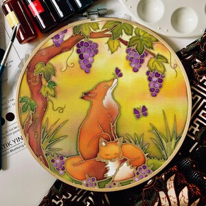 May include: A circular embroidery hoop containing a fabric panel with a hand-painted illustration of two orange foxes under a grapevine. The scene features purple grape clusters, green leaves, and two purple butterflies against a yellow background.