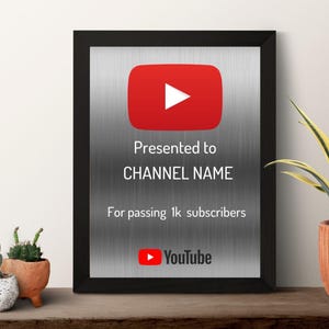 May include: Framed YouTube play button award with a red play button logo. The award is presented to a channel for passing 1k subscribers. The frame is black, and the background is silver. The award is displayed on a wooden shelf with plants.