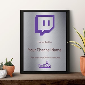 Może przedstawiać: Oprawiona srebrna tablica z fioletowym logo Twitch i tekstem. Tekst brzmi „Prezent dla Nazwy Twojego Kanału Za przekroczenie 1000 subskrybentów”. Tablica znajduje się na drewnianej powierzchni z roślinami doniczkowymi.