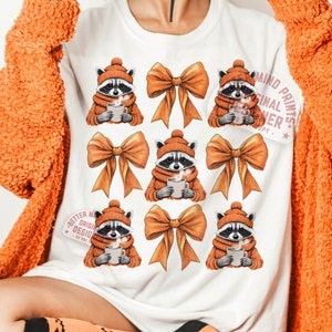 Könnte beinhalten: Weißes T-Shirt mit einem sich wiederholenden Muster aus Waschbären, die orangefarbene Schals tragen und Tassen halten. Jeder Waschbär trägt eine braune Mütze und wird von einer großen orangefarbenen Schleife begleitet. Der Text "Better Mind Original Designs" befindet sich auf der linken Seite des T-Shirts und "Mind Prints Original Designer" auf der rechten Seite des T-Shirts.