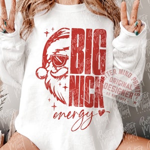 May include: White long-sleeve sweatshirt with a red graphic of Santa Claus wearing sunglasses and the words "BIG NICK energy." The design includes stars and a heart. The sweatshirt is part of a digital download.