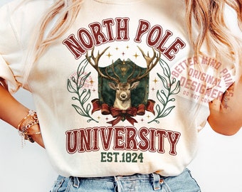 May include: Cream-colored t-shirt featuring a vintage-style graphic. The design includes the words "North Pole University" in a burgundy font, a deer emblem, and the establishment year "EST.1824". The shirt has short sleeves and a relaxed fit.
