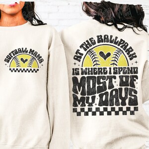 May include: A white sweatshirt with a black and yellow graphic design. The design features a softball with a heart and the text "Softball Mama" on the front. The back of the sweatshirt features the text "At the Ballpark is where I spend most of my days" with a checkered pattern.